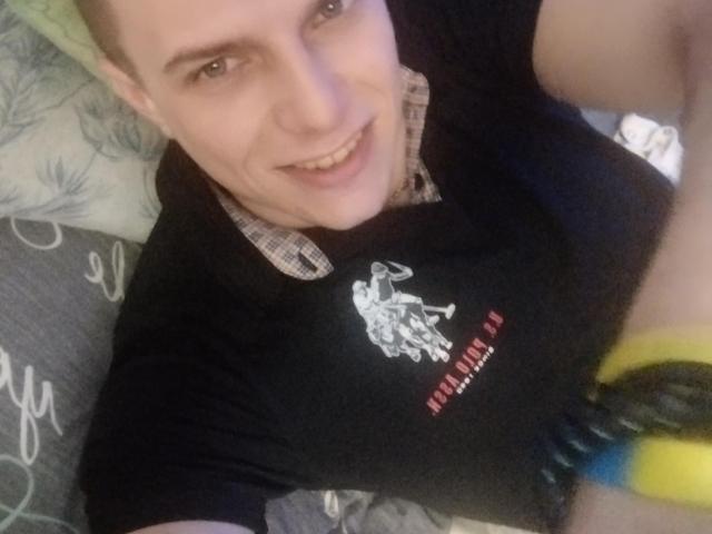 FortuneNorman has a smiling face, light skin, and short brown hair. He's wearing a black shirt with a white graphic and a checkered shirt underneath. His wrist has yellow and black bracelets.