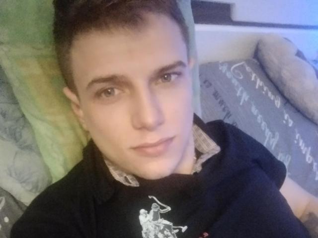 FortuneNorman has short brown hair, blue eyes, and a slight smile. Wearing a black shirt with a white graphic, lying on a green pillow. Soft, relaxed facial expression.