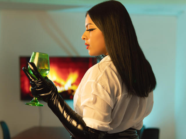 LindaFrancoo with long brown hair, white shirt, black glossy gloves, holding green glass, looking side, lit-up fire in background, sexy, elegant, holding glass, firelight glow.