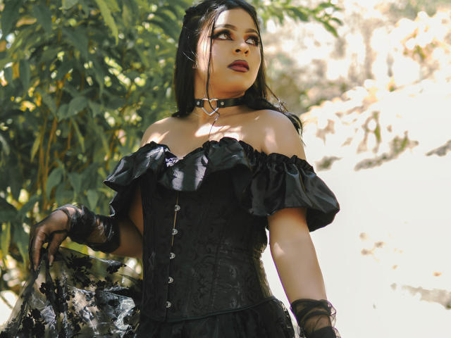 LindaFrancoo with black off-shoulder dress, corset, and choker, looking up, has dark hair, red lipstick, black gloves, holding black lace fabric, sunlight, and green leaves in background.