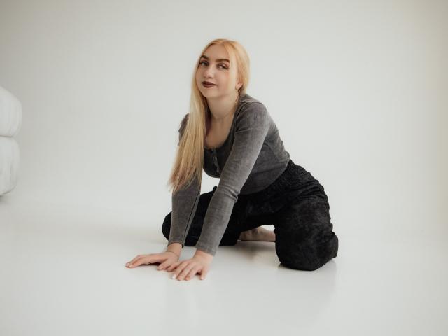 BeatriceFly with long blonde hair, grey long-sleeve top, and black patterned pants, on all fours, looking up with a slight smile. Boobs are small, pussy is not visible.