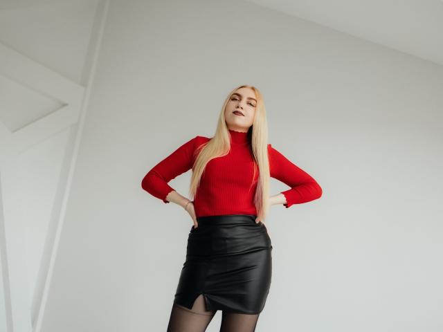 Beatrice, una mujer con cabello rubio largo, un cuello alto rojo, una falda de cuero negro, con las manos en las caderas, de pie en una habitación minimalista. Su trasero está cubierto por la falda. Se ve segura de sí misma..