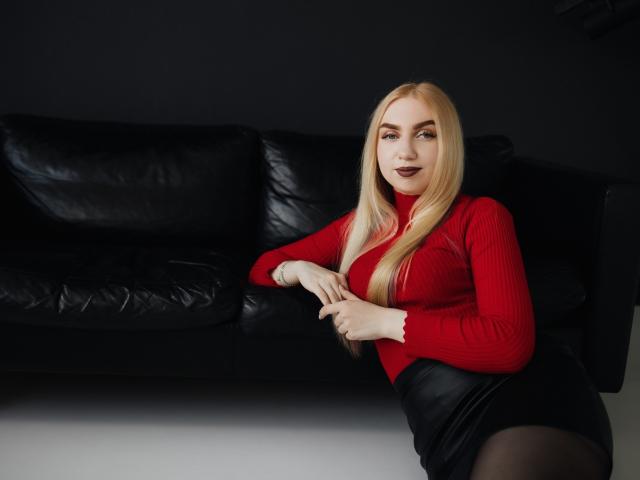 Beatrice, una mujer con cabello rubio largo, un suéter rojo, una falda negra y medias negras, sentada en un sofá de cuero negro, con las manos en las rodillas, sonriendo, con lápiz labial oscuro, sexy, atractiva..