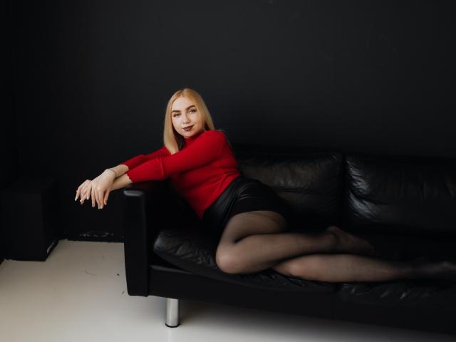 Beatrice, une femme aux cheveux blonds, portant un pull rouge, une jupe noire et des bas noirs, allongée sur un canapé en cuir noir, les jambes croisées, les bras reposant sur le accoudoir, souriante..