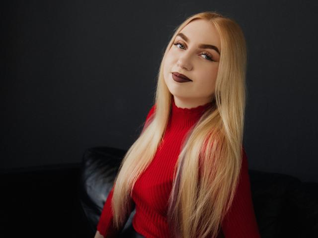 BeatriceFly with long blonde hair, red ribbed sweater, dark lipstick, and slight smile, sitting on black leather couch.