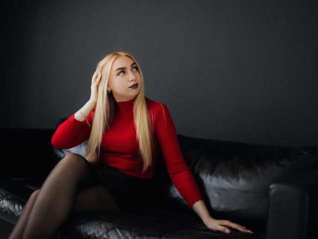 BeatriceFly, rubia, con cabello largo, un cuello alto rojo, una falda negra y medias negras, está sentada en un sofá de cuero negro, con la mano en el pelo, mirando hacia un lado y con lápiz labial oscuro..
