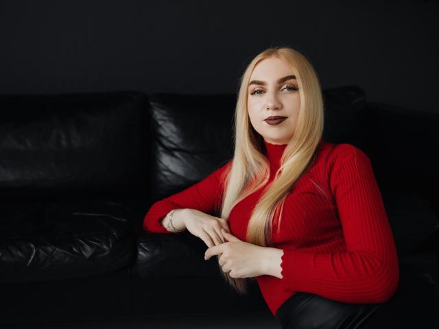 BeatriceFly with long blonde hair, red ribbed sweater, and dark lipstick, sitting on black leather couch, hands resting on lap, looking slightly to the side.