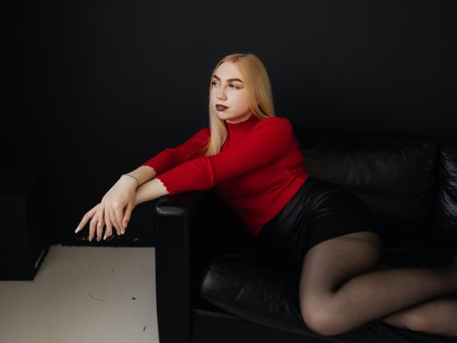 Beatrice, une femme aux cheveux blonds, portant un pull rouge, une jupe noire et des bas noirs, est assise sur un canapé en cuir noir, les bras reposant sur les accoudoirs, les jambes croisées, et porte un rouge à lèvres foncé..