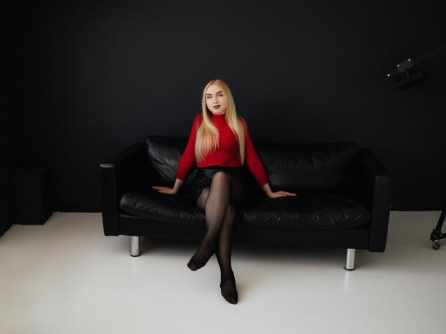 Beatrice: Uma mulher com cabelos loiros longos, vestindo um suéter vermelho, uma saia preta e meias pretas, sentada em um sofá de couro preto, com as pernas cruzadas, usando saltos pretos, sorrindo e com as mãos apoiadas no sofá..