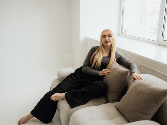 BeatriceFly with blonde hair, gray long-sleeve top, black pants, bare feet, sitting on beige sofa, arms resting on brown pillows, relaxed, smiling, near window, white walls.