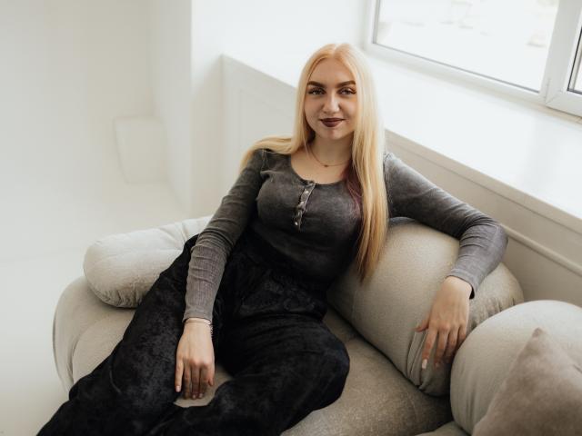 BeatriceFly, une blonde, avec des seins, portant un haut gris à manches longues et un pantalon noir, assise sur un canapé beige, souriant à la caméra, dans une pièce lumineuse avec une fenêtre..
