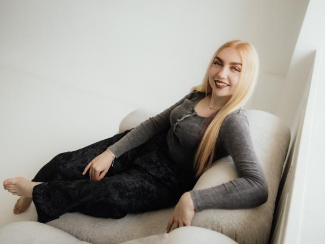 BeatriceFly with blonde hair, gray long-sleeve shirt, and black pants, smiling on a white couch, showing her tits and ass, barefoot, right hand on her thigh.