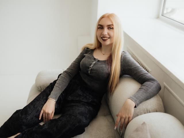 Beatrice, een vrouw met blond haar, een grijze top en zwarte kanten broek, zittend op een witte bank, met een glimlach en donkere lipstick, haar handen rustend op de kussens, in de buurt van een raam..