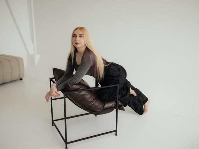 Beatrice: Uma mulher com cabelos loiros longos, blusa cinza, calças pretas estampadas, e uma cadeira de couro marrom, inclinada para frente em uma estrutura de metal preto, mostrando sua nádega e dedos dos pés nus..