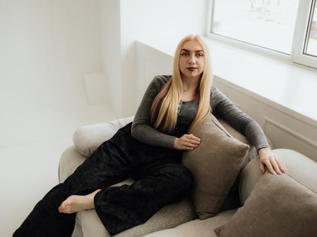 BeatriceFly with long blonde hair, gray long-sleeve shirt, black pants, and bare feet, lounging on a beige sofa with brown pillows, looking relaxed and confident.