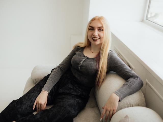 BeatriceFly has long blonde hair, gray top, black lace pants, sitting on a white couch, smiling with dark lipstick, arms resting on cushions, near a window.