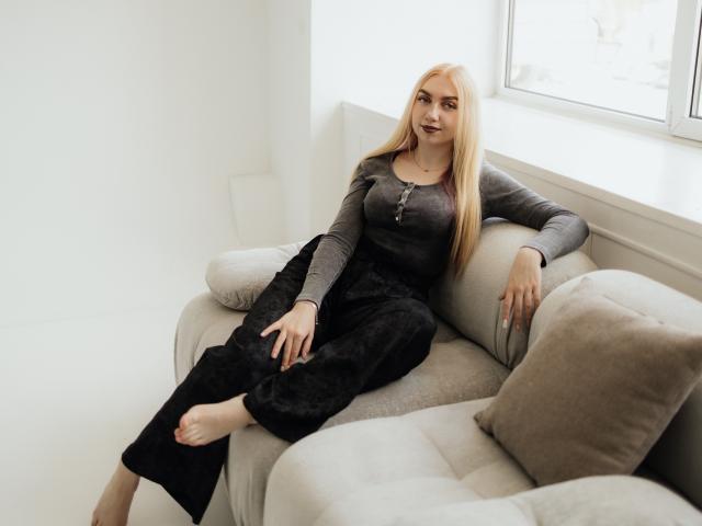 BeatriceFly, loira, com cabelos longos, vestindo uma blusa cinza, calças pretas e descalça, está deitada em um sofá cinza, olhando para a câmera, com um leve sorriso, e um braço apoiado em um travesseiro..