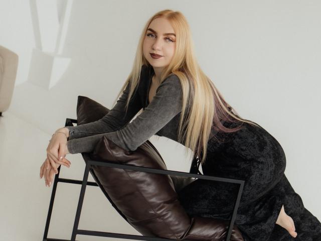 Beatrice, cheveux longs blonds avec des pointes violettes, haut gris, robe noire, appuyée sur une chaise en métal noir, coussin en cuir marron, souriante, rouge à lèvres foncé, fond blanc, pieds nus..