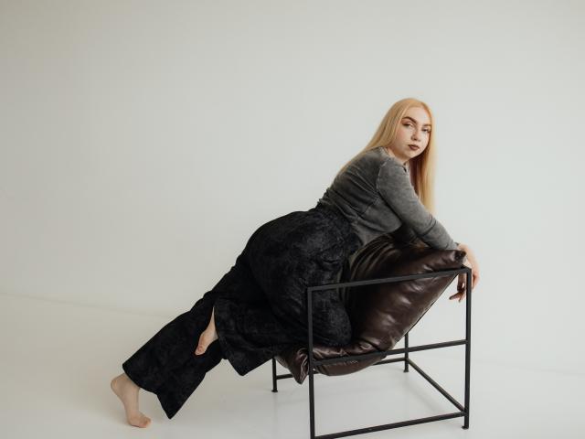 Beatrice, con el pelo rubio largo, una blusa gris y pantalones negros de encaje, apoyada en una silla de cuero marrón, con los pies descalzos sobre el suelo blanco, mirando por encima del hombro..