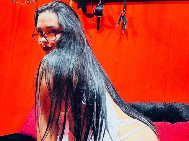CarolaineHotX with long black hair, glasses, and a white lingerie set, showing her back and looking over her shoulder, in a red room with black and red pillows.