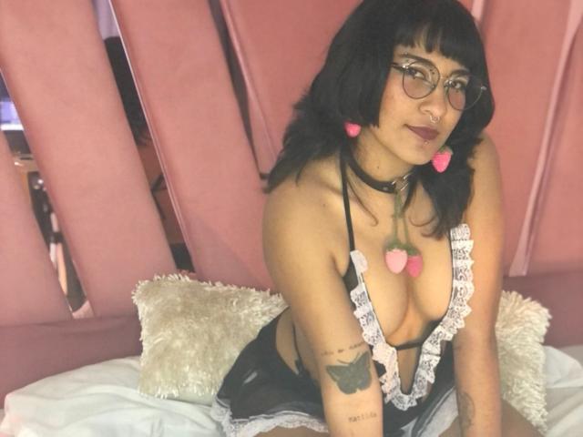FairyHott with black hair, glasses, pink heart earrings, black choker, lace-trimmed top showing big tits, heart necklace, tattoo on left arm, sitting on bed, smiling.