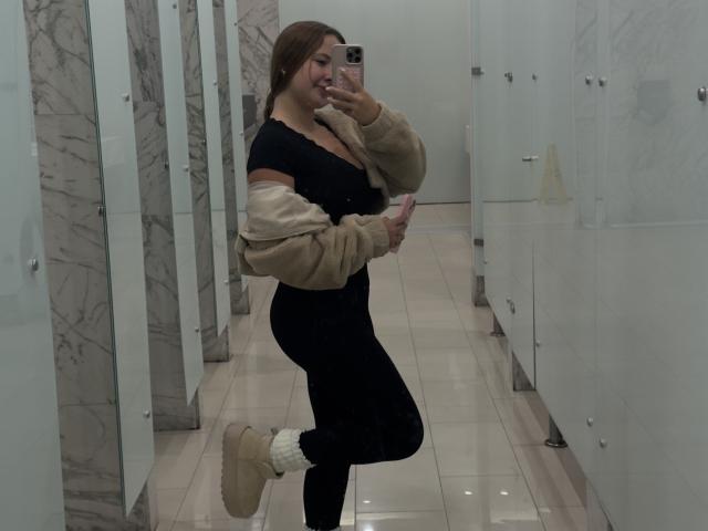 NatSorian, avec ses cheveux bruns, son haut noir, son pantalon noir, sa veste beige et ses chaussettes blanches, prend un selfie devant un miroir dans une salle de bain, montrant ses fesses et souriant..