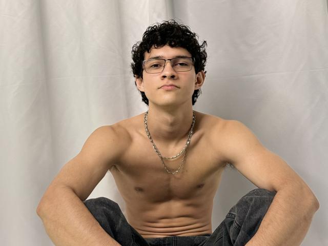 Shirtless SamuelHills with curly black hair, glasses, and a necklace, sitting with legs crossed, showing a toned, light-skinned chest and gray pants.