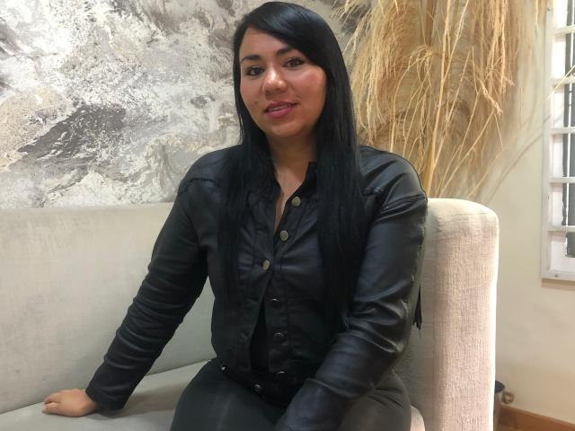 LucyRosse with long black hair, wearing a black leather button-up shirt, sitting on a beige chair, smiling with pink lipstick, and showing a cute ass.