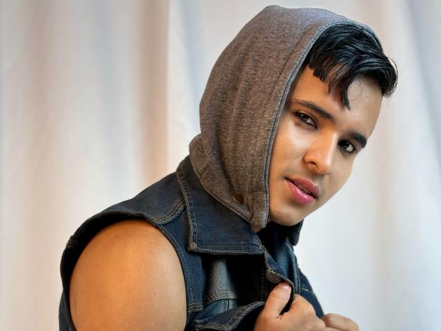 DayanWilly, met kort zwart haar, bruine ogen en lichtbruine huid, draagt een grijze hood en een denim jasje zonder mouwen, en kijkt iets opzij met een subtiele glimlach..