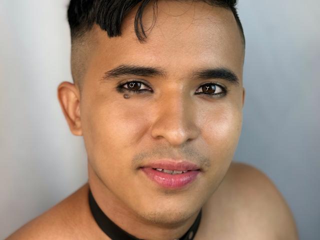 Le visage de DayanWilly, peau bronzée, cheveux noirs courts, yeux marron avec un petit point noir sur le côté gauche, collier noir, lèvres roses, un léger sourire, épaules nues..