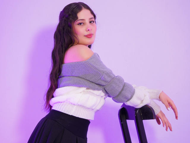 Kathy Stone mit langem, dunklem Haar, grau-weißem Pullover mit Schulterpartie, schwarzem Rock, die an einem schwarzen Stuhl lehnt, violettes Licht, die über die Schulter schaut, sanfter Ausdruck, süß, mittelgroße Brüste..