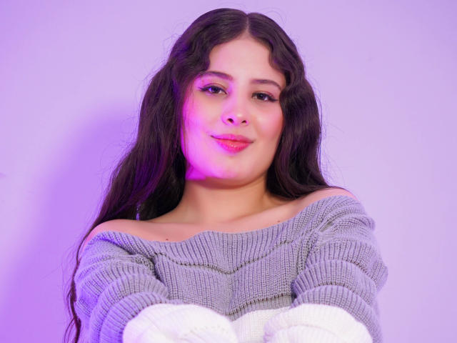 KathyStone with long wavy dark hair, wearing an off-shoulder gray knit sweater, white gloves, and a subtle purple light on her face, smiling gently at the camera.