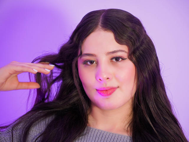 Kathy Stone mit langem, dunklem Haar, pinkem Lippenstift und weißem Nagellack, die ihre Haare hält, trägt einen grauen Pullover, auf einem Foto, das in violettem Licht aufgenommen wurde. Ihr Hintern ist nicht sichtbar..