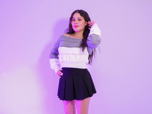 KathyStone with long dark hair, wearing a gray and white striped sweater and black pleated skirt, standing with hand in hair, pink and purple lighting.