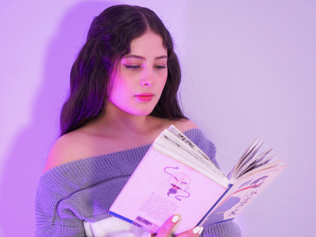 Kathy Stone, con el pelo largo y negro, vestida con un suéter gris de punto con un hombro descubierto, está leyendo un libro y tiene una luz púrpura sobre su rostro. Sus ojos están fijos en el libro..