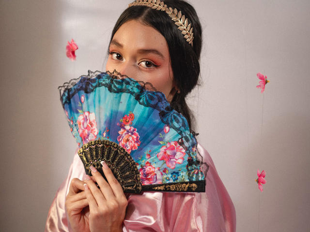 RoseSexAndFun with dark hair, wearing a pink satin robe, gold leaf headband, and blue floral fan covering her tits, pink flower petals floating. Soft pink makeup.