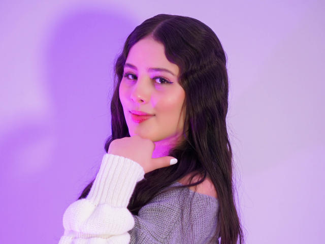 KathyStone with long black hair, wearing a gray off-shoulder sweater and white knitted sleeve, holding her chin with a pink and purple lit background.