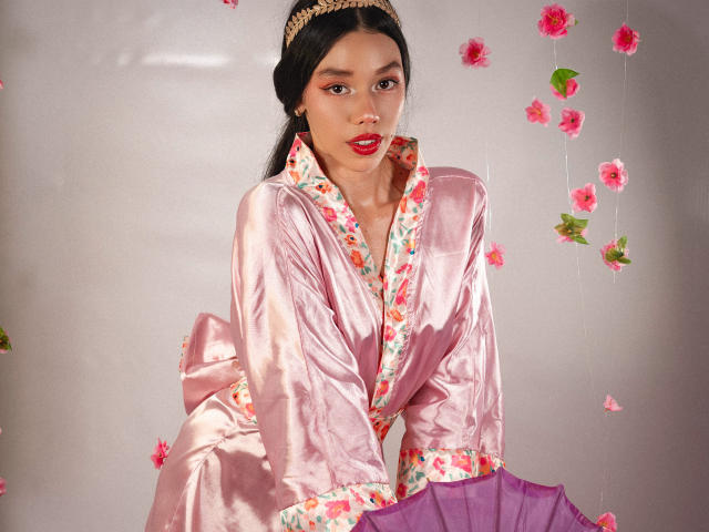 RoseSexAndFun, avec sa robe en satin rose, ses finitions florales, sa jupe violette et son bandeau doré, a des lèvres rouges, des cheveux noirs et des pétales de fleurs roses autour d'elle. Elle est penchée en avant, avec une allure séduisante..