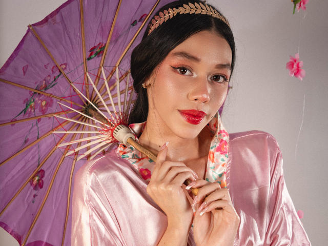 Rose, avec des traits asiatiques, tenant un parapluie floral violet, portant une robe en satin rose et un bandeau orné de feuilles dorées, a du rouge à lèvres et des ongles manucurés, et a une allure mignonne..