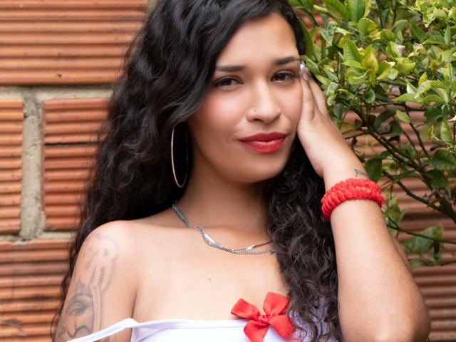 Zoey Moretti, com cabelo preto cacheado, batom vermelho, brincos de argola, corrente de prata, pulseira vermelha, vestido com alça branca, laço vermelho, tatuagem no ombro direito, mão no rosto, sorrindo..