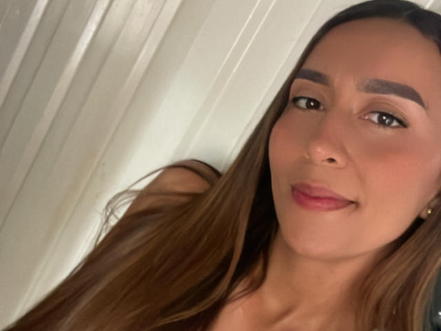 EmelyOwen has long brown hair, brown eyes, and is smiling with pink lips. She's wearing small gold earrings and has well-defined eyebrows. Her face is slightly angled to the right.
