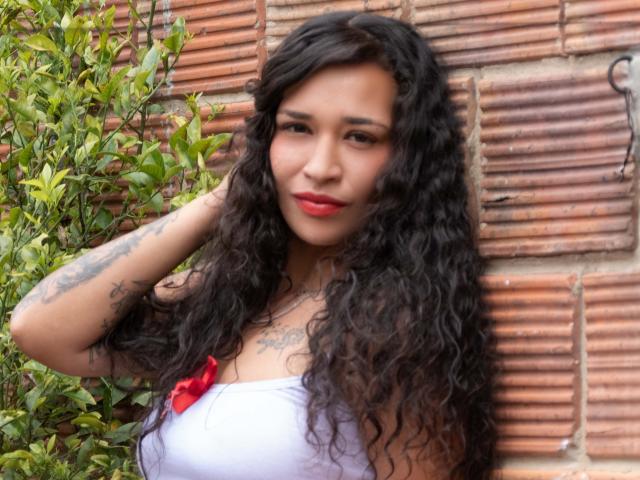 Zoey Moretti mit langen, lockigen, schwarzen Haaren, rotem Lippenstift, einem weißen Top ohne Träger, einem roten Schleifchen, Tattoos an den Armen, steht vor einer Ziegelwand, hat eine Hand im Haar und lächelt. Brüste sind sichtbar..