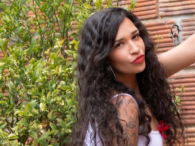 Zoey Moretti, com cabelos pretos longos e cacheados, batom vermelho, camiseta branca, tatuagem visível no braço esquerdo, uma planta verde e uma parede de tijolos vermelhos ao fundo. Suas nádegas não estão visíveis..