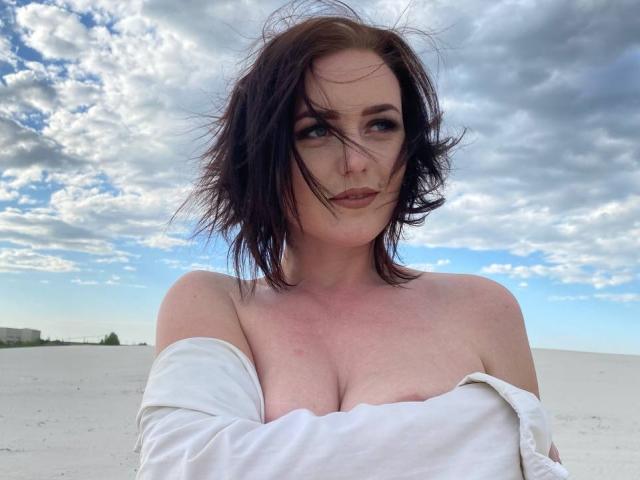 KasandraZ69 with messy dark brown hair, pale skin, and small boobs, wearing a white off-shoulder top, standing in a sandy landscape under a cloudy blue sky.