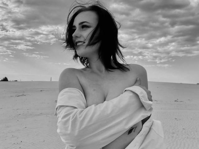 Black-and-white photo of KasandraZ69 with shoulder-length dark hair, smiling, off-shoulder white shirt exposing her tits, arms crossed, standing in a sandy desert with cloudy sky.