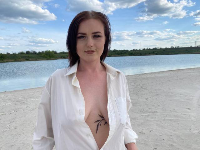 KasandraZ69 with pale skin, brown hair, open white shirt revealing small tits and a black leaf tattoo on her chest, standing on a sandy shore with a calm water background.