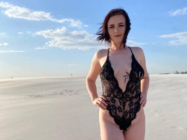 KasandraZ69 stands in a sandy desert wearing a black lace one-piece that reveals her tits and shows her pussy through the sheer fabric. She has dark hair and a confident expression.