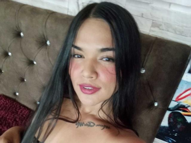 Samanta Moss, con i suoi lunghi capelli neri, le labbra rosa e un tatuaggio sul petto, è sdraiata su una testiera marrone imbottita, mostrando le spalle scoperte e guardando verso la fotocamera..