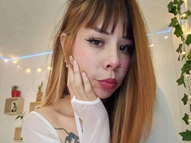 AriiannaHot with light brown hair, nose piercing, and white sheer top, touching her cheek with pink lipstick and black eyeliner, showing a tattoo on her right shoulder.