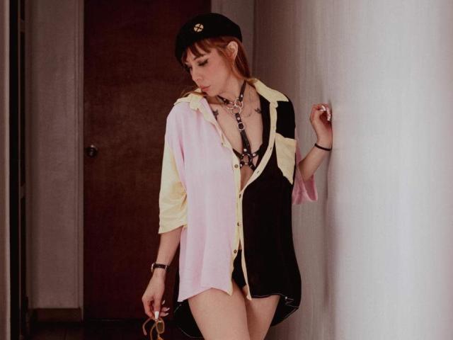 AriiannaHot stands in a hallway, wearing a black-and-pink shirt with a yellow collar, black hat, and necklace, holding glasses in her right hand, showing her tits.